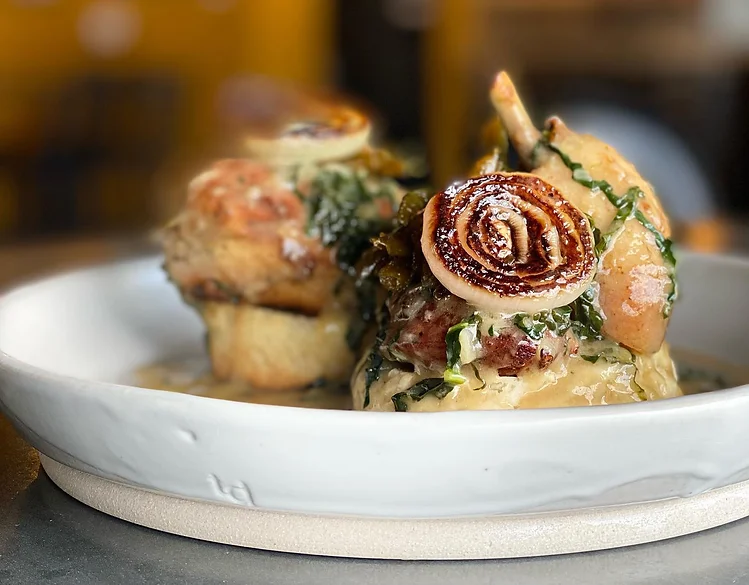 Close-up of a plated dish with biscuits, braised meat, leafy greens, and rolled seared garnish in warm golden tones