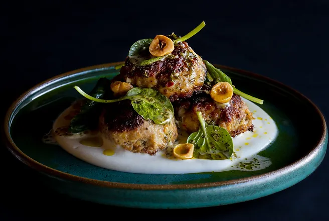 Close-up of stacked golden-brown fried patties topped with greens and nuts on white sauce, served on a teal plate